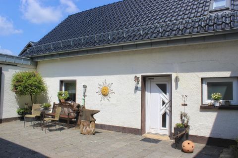 Eine ruhige Terrasse mit Holzbank und Tisch, umgeben von Pflanzen. Das Haus hat eine helle Fassade und ein sonniges Wandschild.