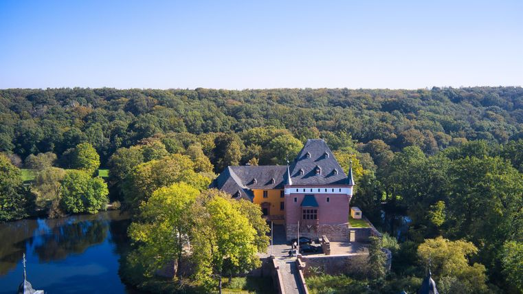 Eine Aussicht auf ein malerisches Schloss umgeben von Bäumen und Wasser. Der klare Himmel ergänzt die serene Landschaft.