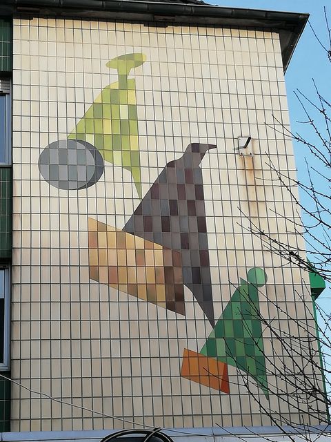 Eine Wandmalerei mit geometrischen Formen in verschiedenen Farben. Die Gestaltung umfasst grüne, braune und graue Elemente auf einem hellen Hintergrund.
