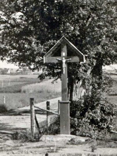 Ein Holzwegkreuz steht an einem ländlichen Weg, umgeben von Bäumen und Wiesen. Im Hintergrund erstreckt sich eine offene Landschaft.