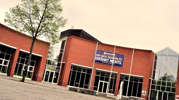 Ein modernes Gebäude mit großen Fenstern und einer einladenden Fassade. Vor dem Eingang steht ein Schild mit der Aufschrift: "Wer nicht neugierig ist, erfährt nichts."