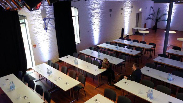 Ein eleganter Veranstaltungsraum mit langen Tischen und Stühlen. Sanftes Licht beleuchtet das Zimmer, das für eine Präsentation oder ein Meeting vorbereitet ist.