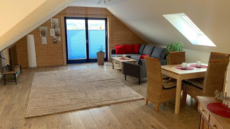 Ein helles, modernes Wohnzimmer im Dachgeschoss mit einem gemütlichen Sofa, einem Tisch und Stühlen. Große Fenster bieten viel Tageslicht und einen Blick nach draußen.