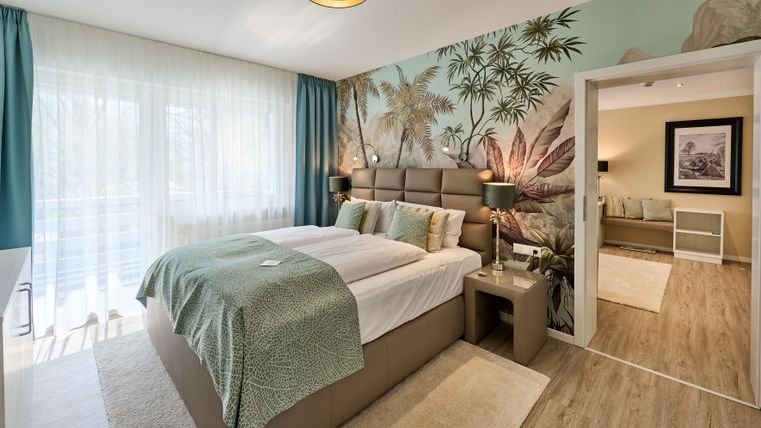 Ein modernes Schlafzimmer mit einem großen Bett und einer stilvollen Wandgestaltung. Viel Licht von den Fenstern sorgt für eine einladende Atmosphäre.