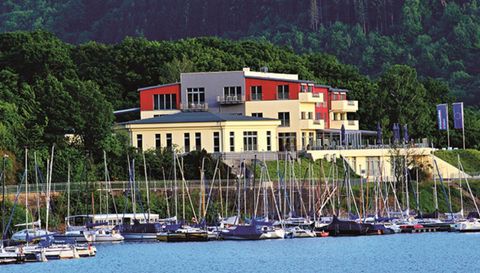 Ein modernes Gebäude am Wasser mit vielen Segelbooten im Vordergrund. Die Umgebung ist grün und bietet eine schöne Aussicht auf die Natur.