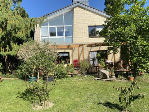 Ein gemütliches Haus mit großen Fenstern und einer überdachten Veranda. Der Garten ist grün mit verschiedenen Pflanzen und Bäumen.