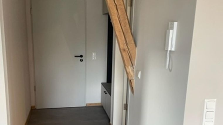 Ein schmaler Flur mit einer weißen Tür und einem Holzstamm an der Wand. Der Boden ist mit Teppich ausgelegt und die Wände sind hell gestrichen.