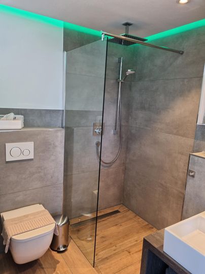 Ein modernes Badezimmer mit einer begehbaren Dusche und eleganten Fliesen. Die Beleuchtung in Grün sorgt für eine angenehme Atmosphäre.