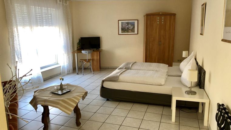 Ein einfaches, helles Zimmer mit einem Doppelbett und einem kleinen Tisch. Es gibt einen Fernseher und große Fenster, die viel Licht hereinlassen.