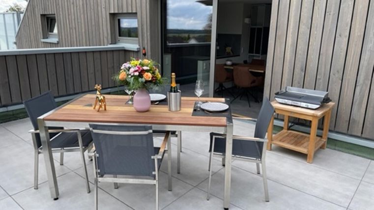 Ein schöner Balkon mit einem Tisch und vier Stühlen. Auf dem Tisch stehen Blumen und eine Flasche, im Hintergrund sind große Fenster zu sehen.