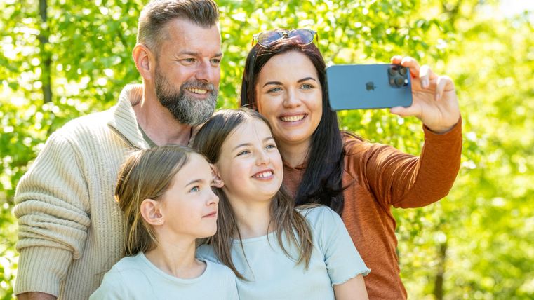 Eine glückliche Familie macht ein Selfie im Freien. Im Hintergrund sind grüne Bäume und viel Sonnenlicht zu sehen.