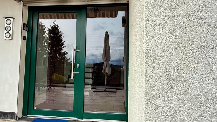 Eine Eingangstür mit einer grünen Rahmung und einem blauen Fußmatte. Im Hintergrund sind Bäume und ein Sonnenschirm sichtbar.