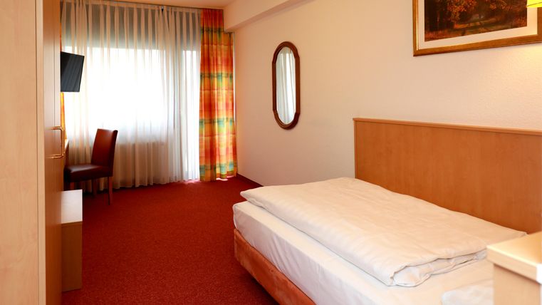 Ein einfaches Hotelzimmer mit einem Bett, einem Tisch und einem Stuhl. Der Raum hat einen Teppichboden und große Fenster mit Vorhängen.