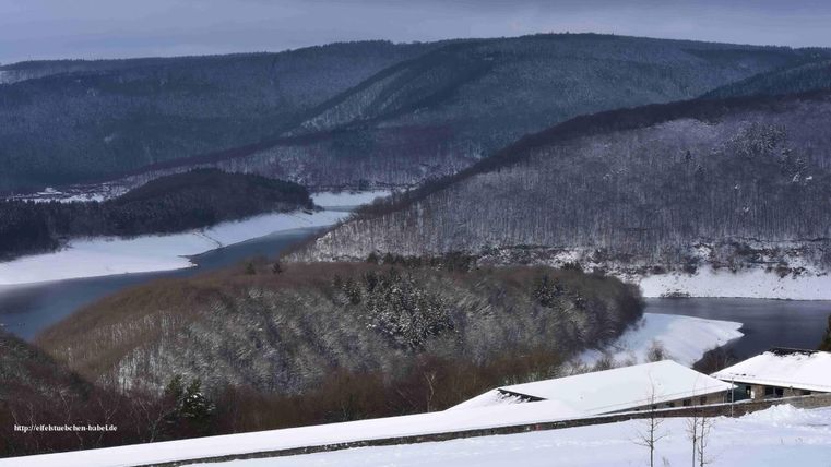 Eine winterliche Landschaft mit schneebedeckten Hügeln und einem glitzernden Fluss. Der Himmel ist bewölkt und verleiht der Szene eine ruhige Atmosphäre.