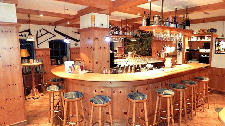 Eine gemütliche Bar mit Holzdekor, an der mehrere Personen stehen und sitzen. Es gibt freundliche Gespräche und Getränke werden serviert.