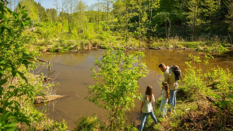 Eine Familie steht an einem ruhigen Gewässer umgeben von üppigem Grün. Die Sonne scheint und es ist ein schöner Tag in der Natur.