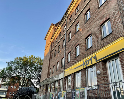 Ein graues Backsteingebäude mit einem gelben Schild. Im Vordergrund sind Bäume und ein Bereich mit Plakaten zu sehen.