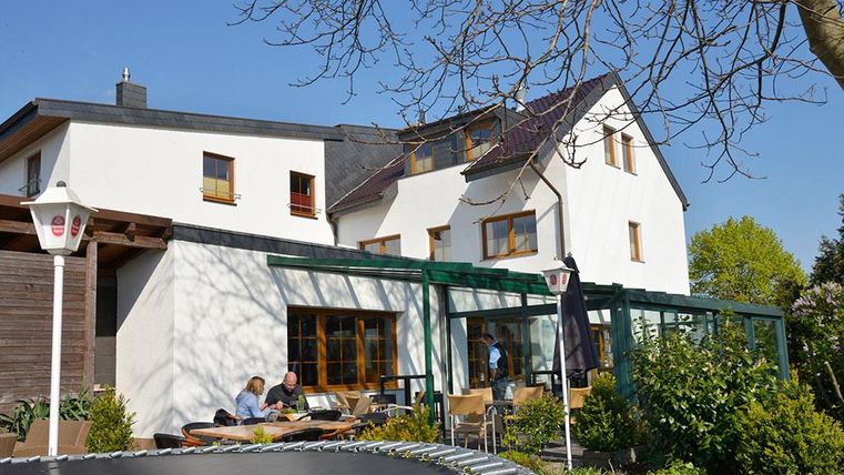 Ein modernes Haus mit einem Garten und einer Terrasse. Menschen sitzen draußen und genießen die Sonne.