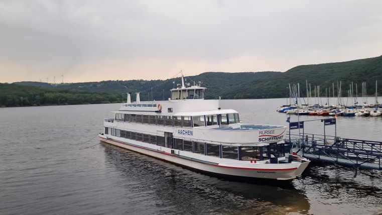 Ein Ausflugsboot liegt am Steg im Wasser. Im Hintergrund sind sanfte Hügel und ein Yachthafen zu sehen.