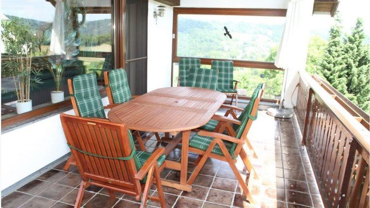 Ein gemütlicher Balkon mit einem Holzstuhlset und einem Tisch. Im Hintergrund sind Bäume und eine schöne Aussicht zu sehen.