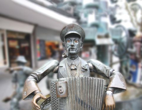 Eine bronzene Statue eines Mannes, der ein Akkordeon spielt. Im Hintergrund sind unscharfe Geschäfte zu sehen.