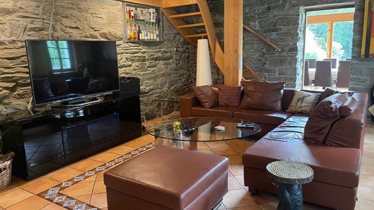 Ein gemütliches Wohnzimmer mit einem großen Fernseher und einer braunen Ledergarnitur. Das Holzdecke und die Steinwände verleihen dem Raum einen rustikalen Charme.