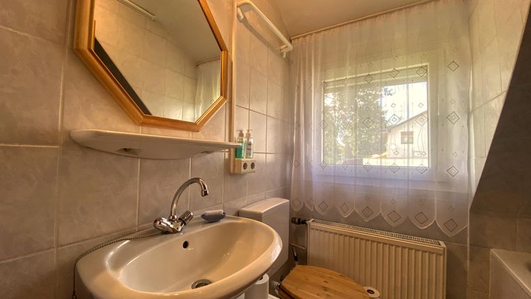 Ein kleines Badezimmer mit einem Waschbecken, einer Toilette und einem Fenster. Die Wände sind hell gefliest und ein runder Spiegel hängt über dem Waschbecken.