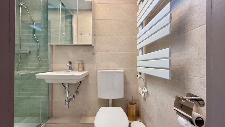 Ein modernes Badezimmer mit einer Dusche, einem Waschbecken und einer Toilette. Die Wände sind hell und mit fliesenähnlichem Material gestaltet.