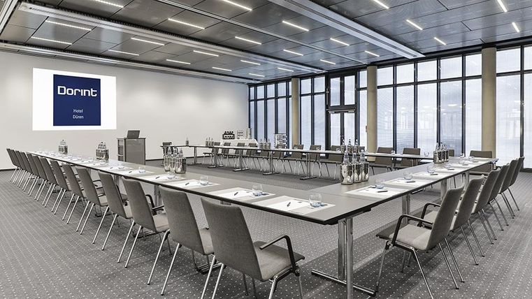 Ein moderner Konferenzraum mit u-förmiger Tischanordnung. Große Fenster sorgen für viel Tageslicht und eine professionelle Atmosphäre.