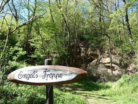 Ein Wegweiser mit der Aufschrift „Engel’s Tropp“ steht vor einem bewaldeten Bereich. Die Umgebung ist grün und üppig, mit Bäumen und einem Felsen im Hintergrund.