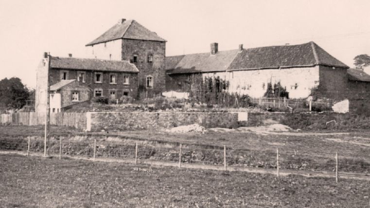 Ein historisches Bauernhaus mit einem großen, angelegten Garten. Im Hintergrund sind weitere Gebäude sichtbar, umgeben von einer ländlichen Umgebung.
