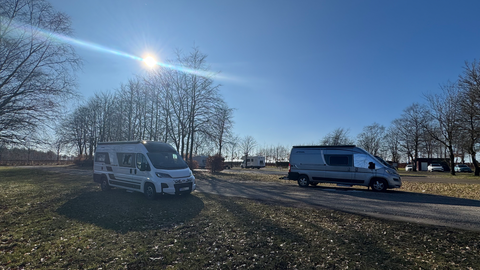 Zwei Wohnmobile stehen auf einem Platz mit Bäumen im Hintergrund. Der Himmel ist klar und sonnig.