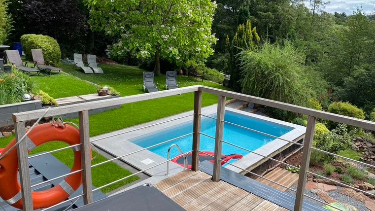 Ein schöner Garten mit einem Pool und grüner Rasenfläche. Im Hintergrund stehen Liegen unter schattenspendenden Bäumen.