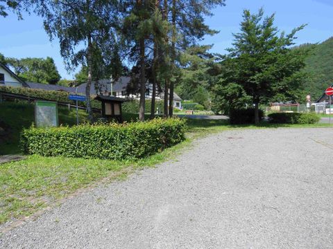 Eine ruhige Straße mit Kiesbelag, umgeben von Bäumen und Büschen. Im Hintergrund sind einige Gebäude und ein Spielplatz zu sehen.