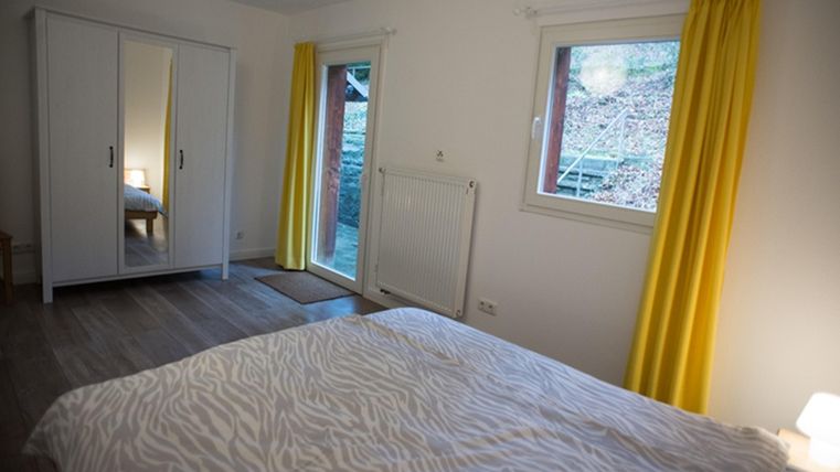 Ein helles Schlafzimmer mit einem Bett und gelben Vorhängen. Der Raum hat einen Kleiderschrank und Fenster, die auf einen Garten blicken.