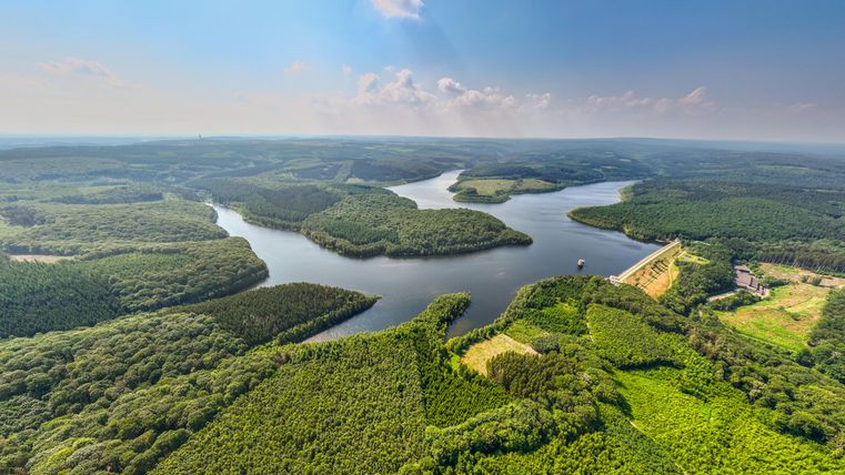 Zwei Wanderer gehen einen schmalen Pfad entlang eines Baches im Wald. Die Sonne scheint und um sie herum sind grüne Bäume und trockenes Laub.