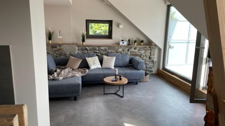 Ein modernes Wohnzimmer mit einem grauen Sofa und einem Couchtisch. Große Fenster lassen viel Tageslicht herein und bieten einen Blick nach draußen.