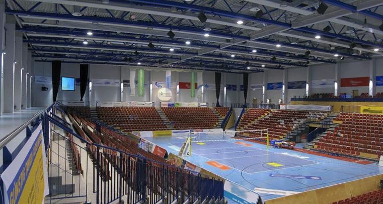 Eine große Sporthalle mit blauen Boden und vielen roten Sitzplätzen. Im Hintergrund sind Tribünen und Werbebanner sichtbar.
