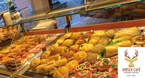Eine Auswahl an köstlichen Backwaren und Snacks in einer Glasvitrine. Im Hintergrund sind frische Brötchen und verschiedene belegte Brote zu sehen.