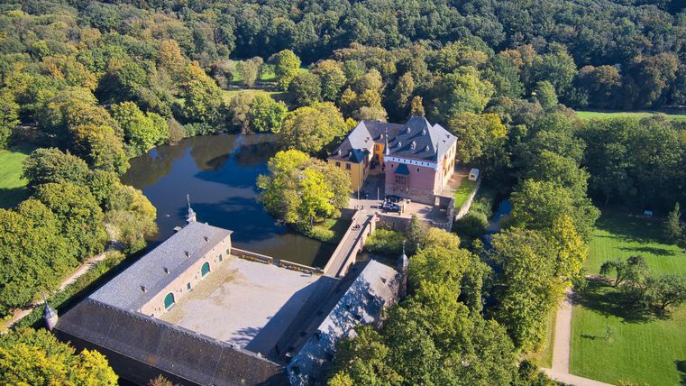 Eine malerische Ansicht von einem Schloss, umgeben von Bäumen und einem ruhigen Teich. Die Anlage liegt in einer grünen Landschaft und strahlt eine friedliche Atmosphäre aus.