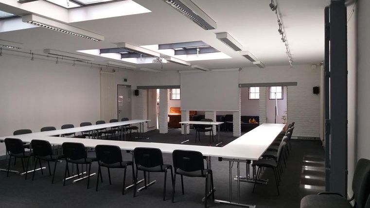 Ein heller Konferenzraum mit langen Tischen und Stühlen. Große Fenster sorgen für viel natürliches Licht.