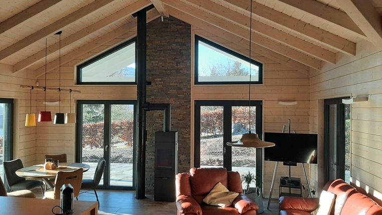 Ein modernes Wohnzimmer mit Holzbalken und großen Fenstern. Es gibt gemütliche Möbel und einen Kamin, der eine einladende Atmosphäre schafft.