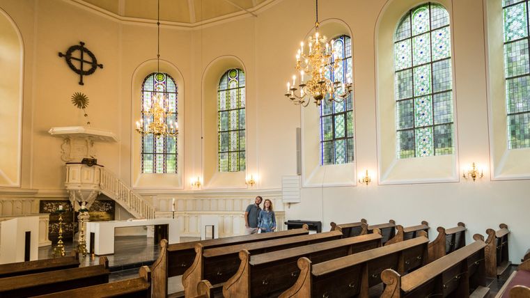 Eine ruhige Kirche mit Holzbankreihen und bunten Fenstern. Im Vordergrund stehen zwei Personen, die den Raum betrachten.