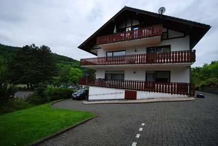 Ein modernes Gebäude mit mehreren Etagen und Balkonen befindet sich in einer grünen Umgebung. Die Auffahrt ist gepflastert und von Bäumen umgeben.