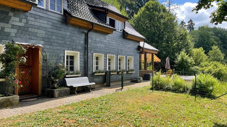 Ein charmantes, graues Haus mit einem schönen Garten. Im Hintergrund sind Bäume und eine sonnige Landschaft zu sehen.