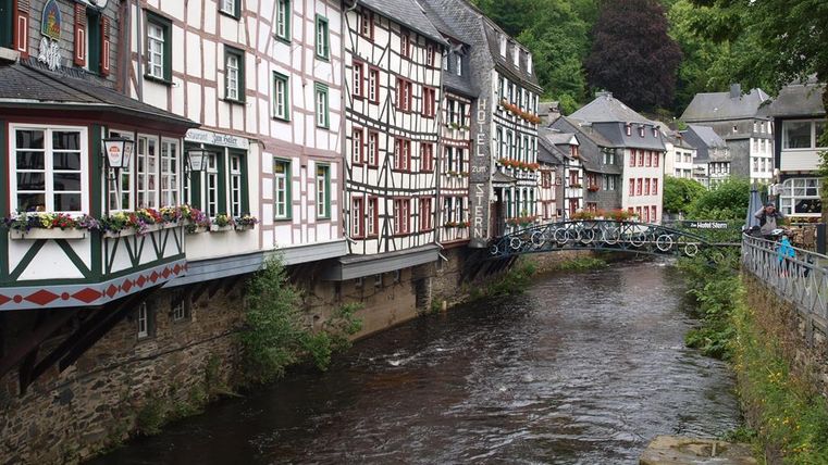 Eine malerische Stadtansicht mit Fachwerkhäusern entlang eines ruhigen Flusses. Die grüne Landschaft im Hintergrund ergänzt die charmante Atmosphäre.