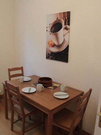 Ein kleiner Essbereich mit einem Holztisch und zwei Stühlen. An der Wand hängt ein Bild von einer Kaffeetasse mit einem Keks.