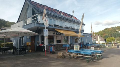 Ein Kioskgebäude mit Außenterrasse und bunten Tischen. Im Hintergrund sind grüne Hügel und eine klare Himmel sichtbar.