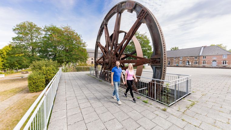 Ein großes historisches Rad steht in einem Park. Zwei Personen spazieren entspannt auf dem Gehweg neben dem Rad.