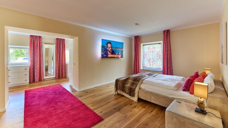 Ein modernes Schlafzimmer mit einem großen Bett und Fernseher. Der Raum hat Holzböden und einen roten Teppich, sowie Vorhänge in sanften Farben.